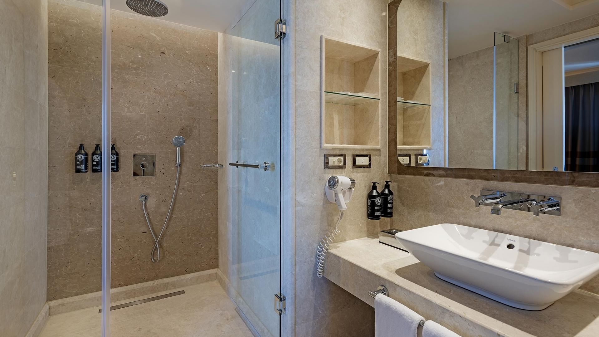 Titanic City Taksim'deki deluxe odanın banyosunda cam duşakabin, beyaz lavabo, havlu ve şık detaylar görülüyor.