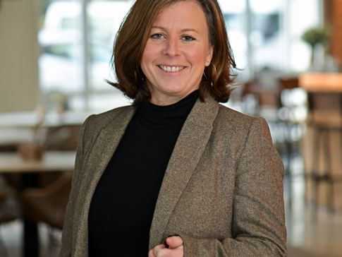 Femme souriante en blazer brun et haut noir à l'Hôtel Rivella.