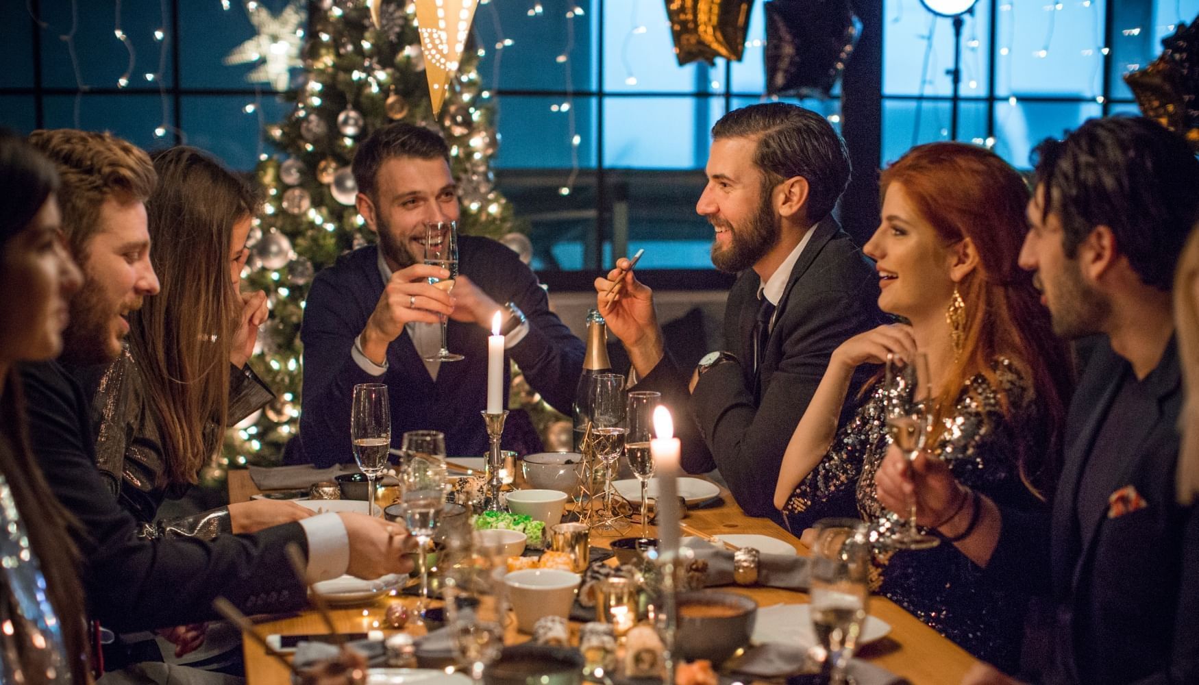 Group of people seated around a dining table with food, candles, and drinks, celebrating in front of a Christmas tree.