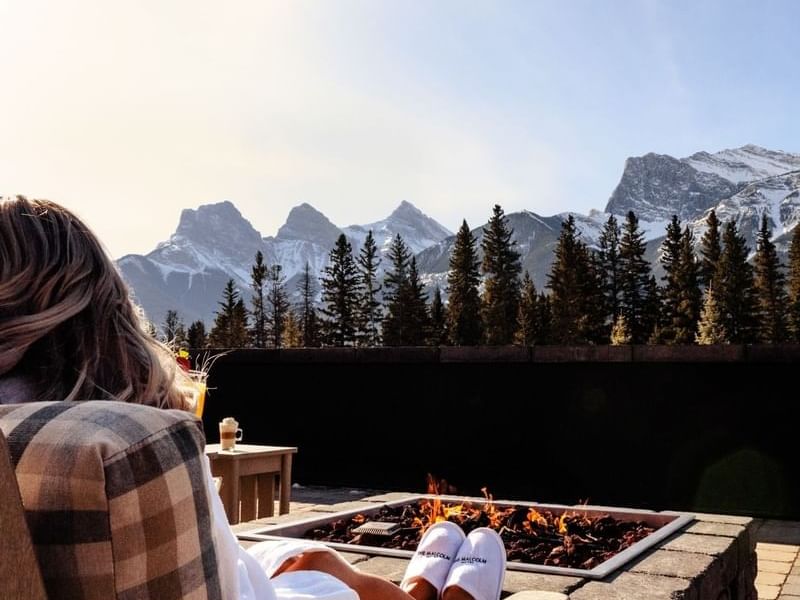 Woman in white robe and slippers relaxing on a bench by a fire pit with mountain view.