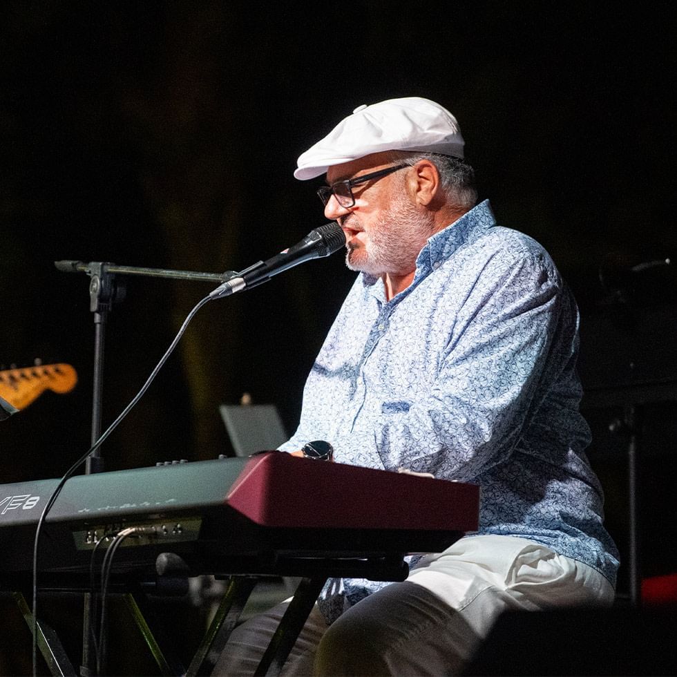 Ein Mann mit Hut und Brille spielt Keyboard und singt in ein Mikrofon auf einer Bühne.