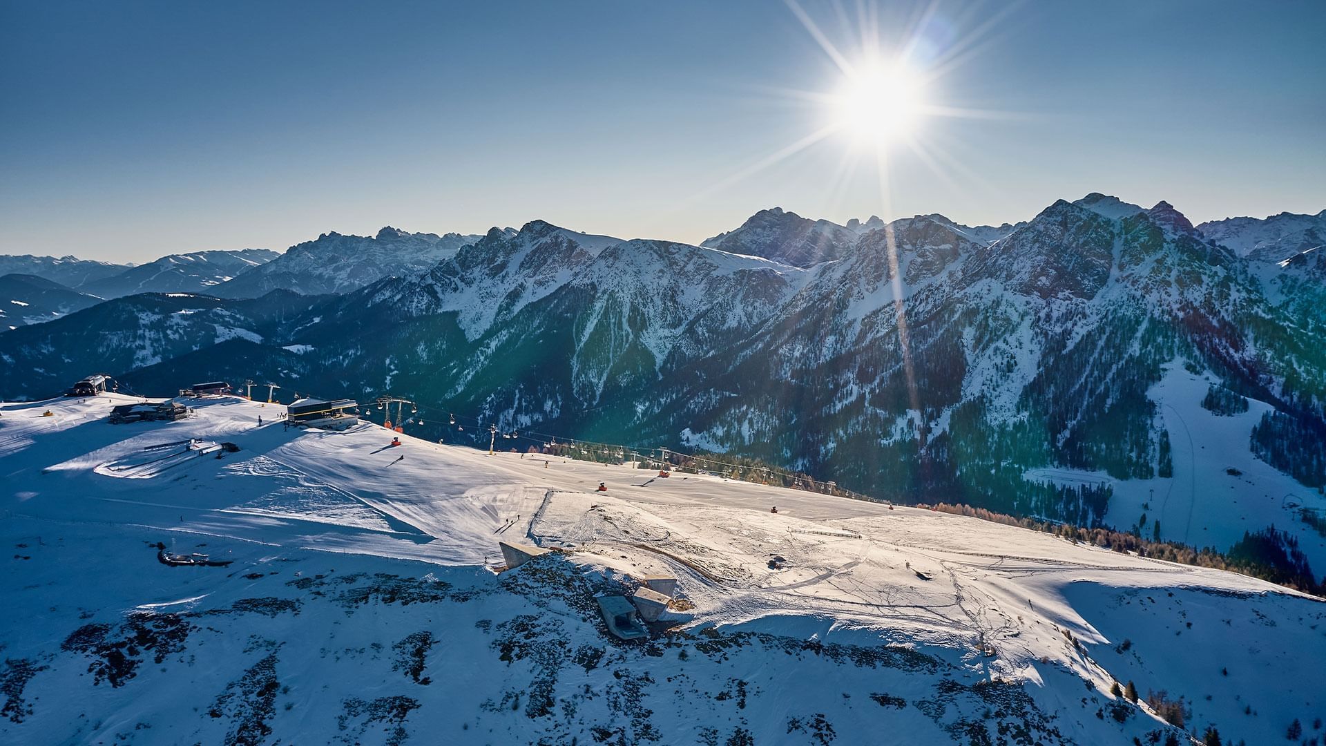 Panorama delle Alpi innevate con impianti di risalita, sotto un cielo azzurro e un sole luminoso.