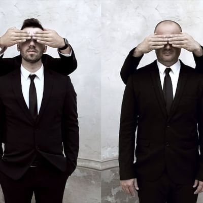 Zwei Männer in schwarzen Anzügen und Krawatten, die sich gegenseitig die Augen bedecken.