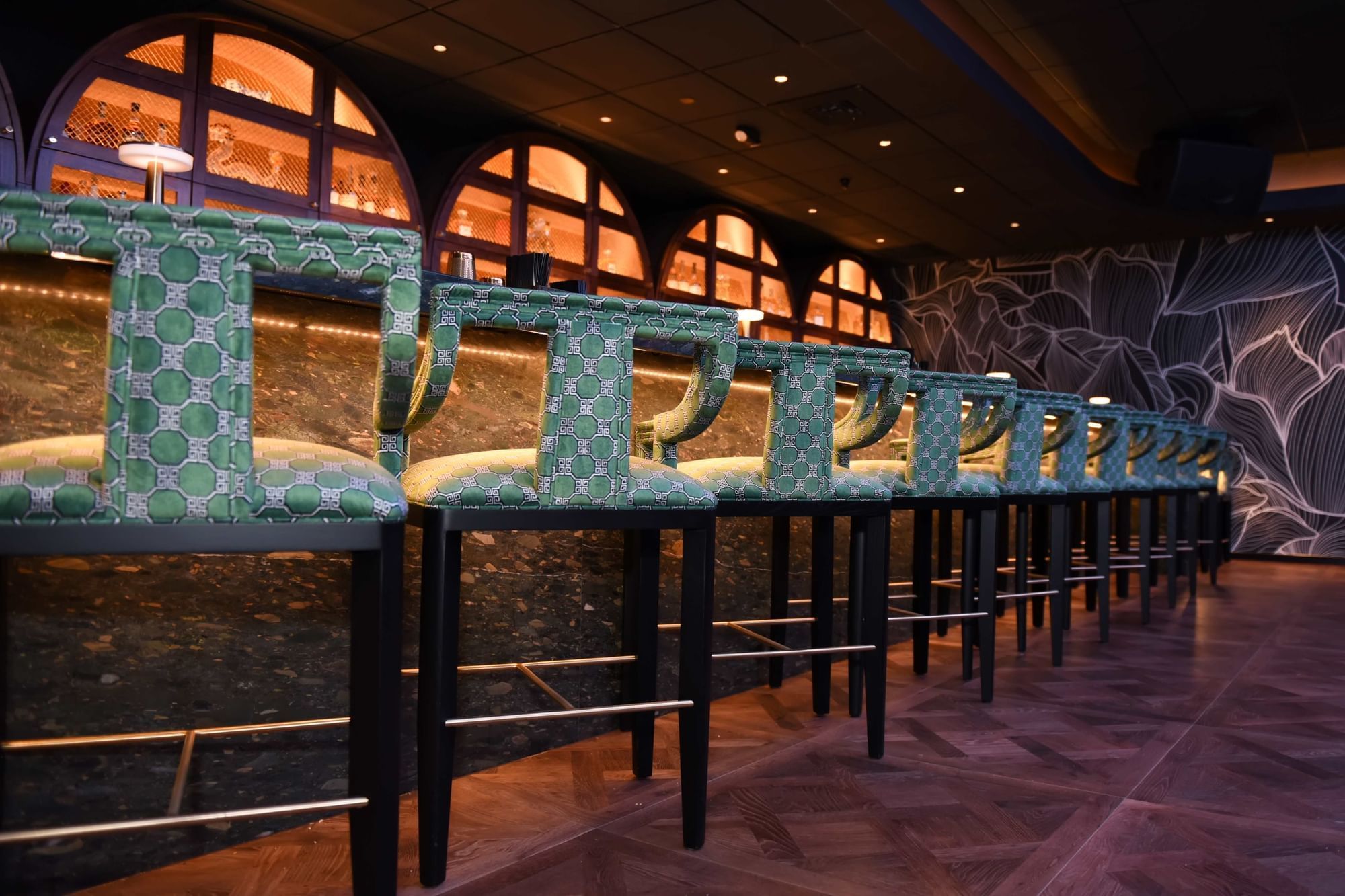 Green patterned bar stools lined by a stone counter under arched bottle displays and warm lights at The Brook