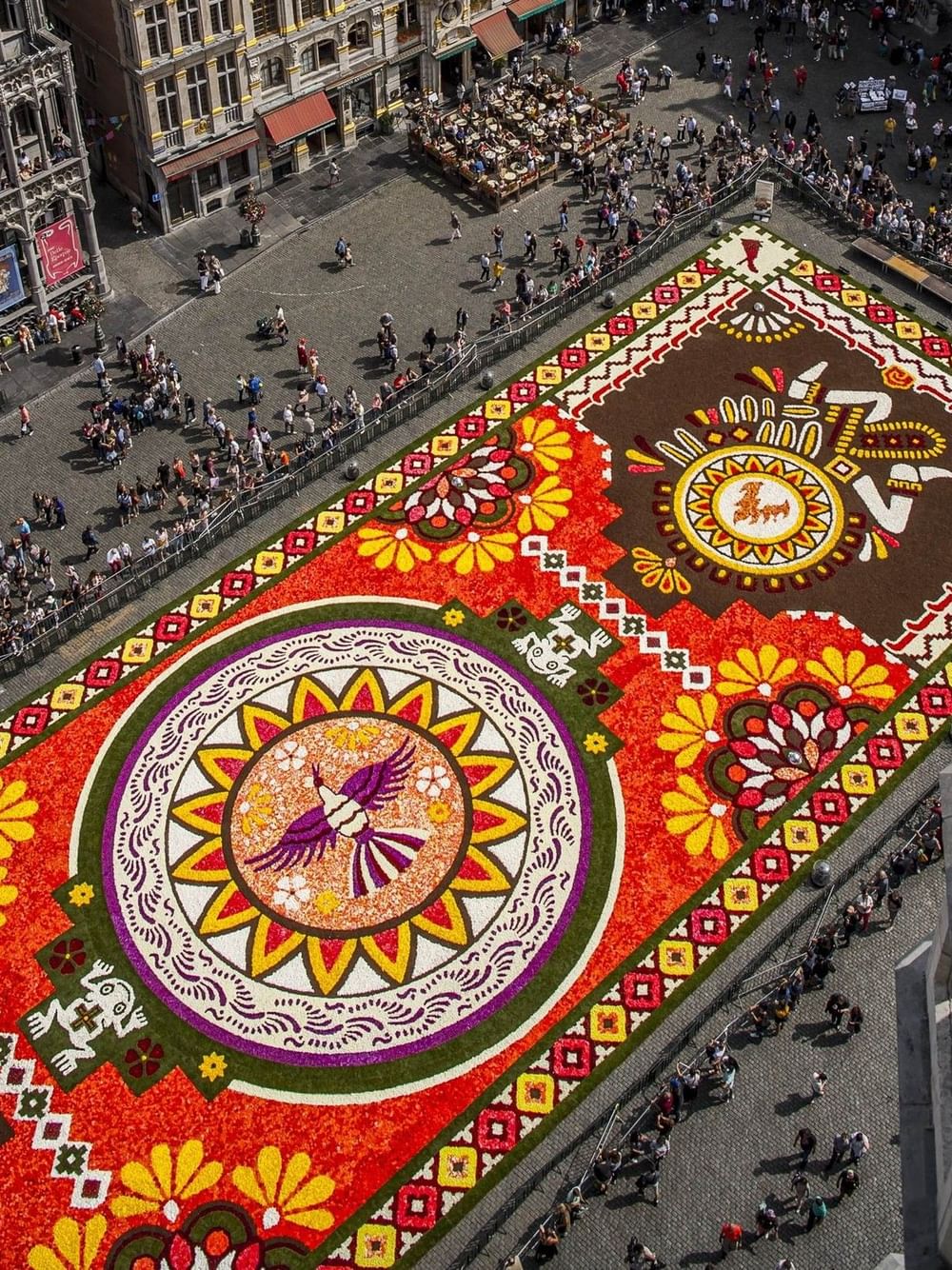 Luchtfoto van een kleurrijk bloementapijt met geometrische patronen op een historisch stadsplein omringd door bezoekers.