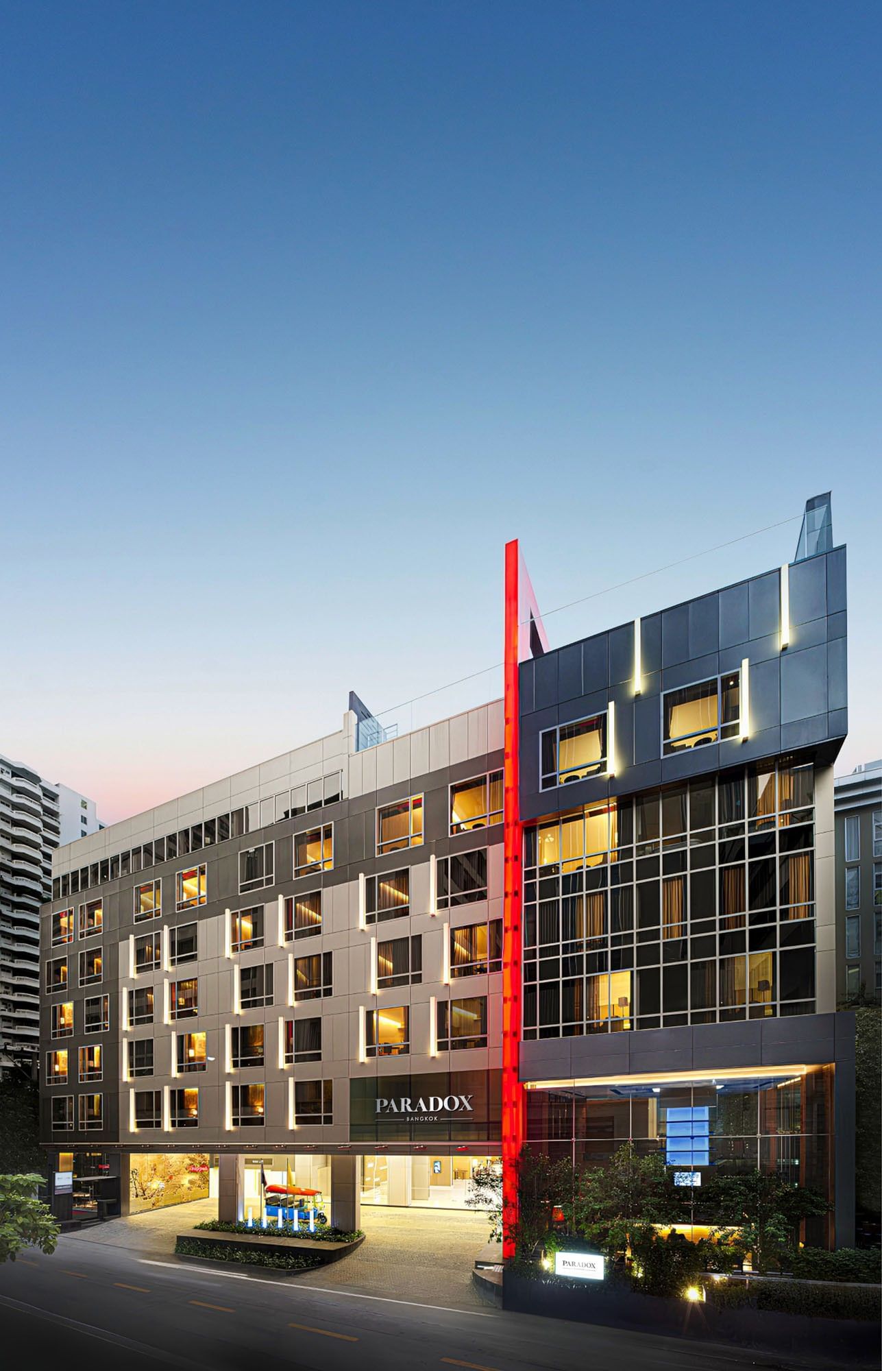 Modern facade of Paradox Bangkok Hotel on Sukhumvit Road, featuring a red stripe by lit windows under a clear evening sky