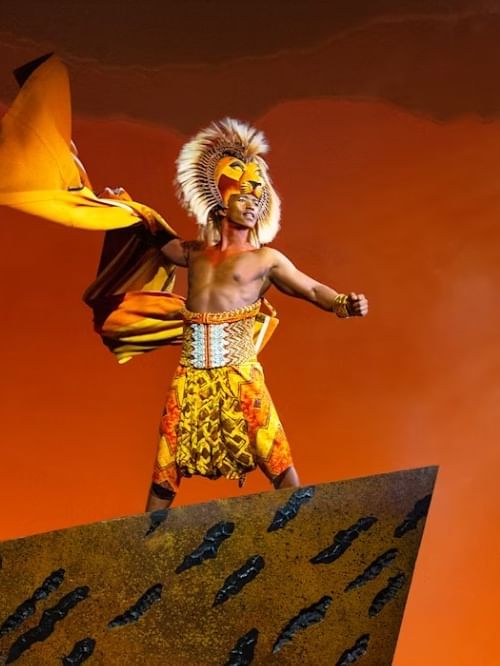 Performer in lion costume and headdress stands on Pride Rock against orange sky in Disney's THE LION KING production.