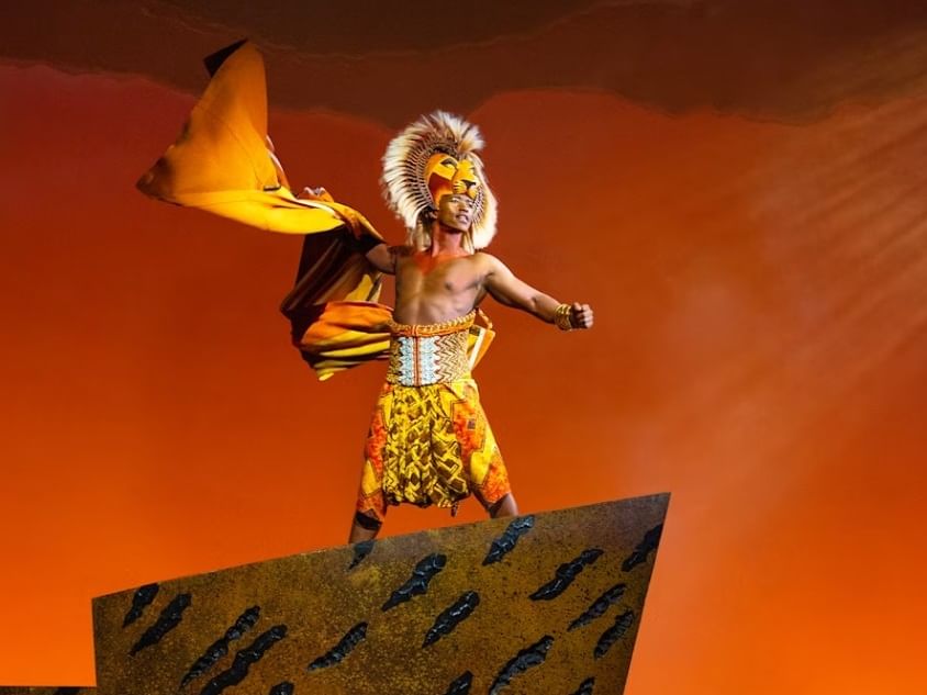 Performer in lion costume and headdress stands on Pride Rock against orange sky in Disney's THE LION KING production.