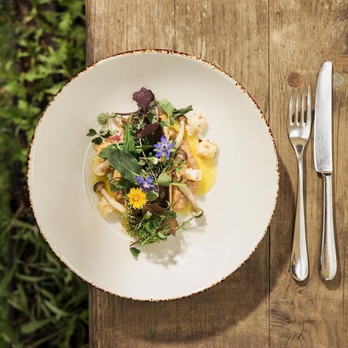 Plate of salad with herbs and flowers, knife and fork on a wooden table.