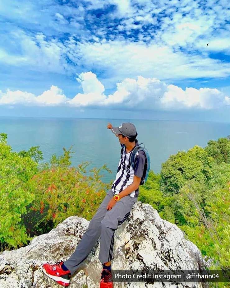 Hiker sitting on a rocky cliff pointing at the ocean view - Lexis Port Dickson