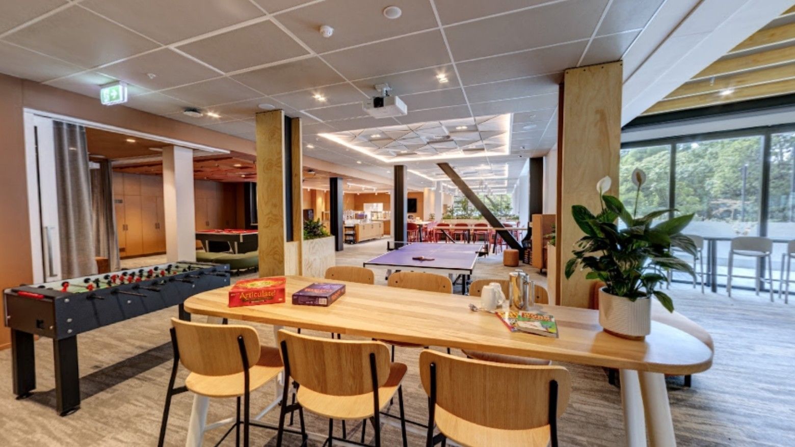 Modern hotel recreation area with foosball table, wooden communal tables, board games, and natural light from large windows