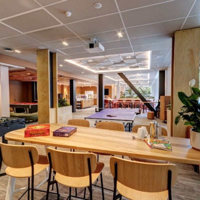 Modern hotel recreation area with foosball table, wooden communal tables, board games, and natural light from large windows