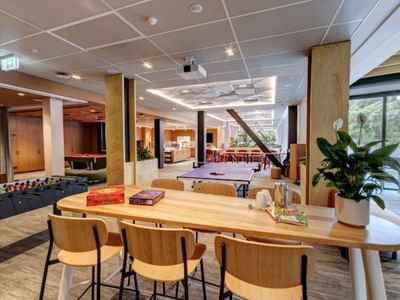 Modern hotel recreation area with foosball table, wooden communal tables, board games, and natural light from large windows