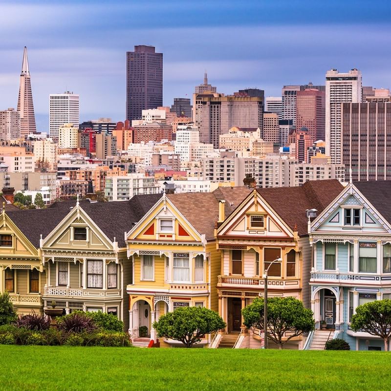 Colorful Victorian houses line a green lawn in front of the San Francisco skyline near Warwick Hotels and Resorts