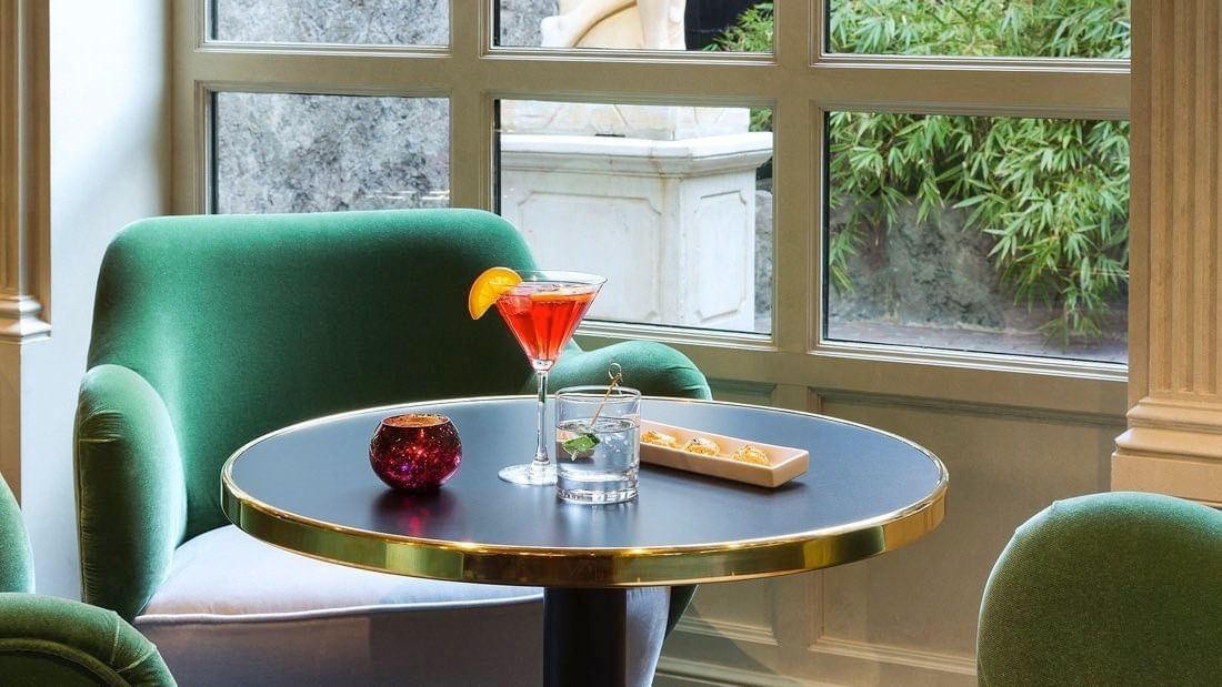 Green chairs around a table with a cocktail, water, and snacks near a window at Hotel Barsey by Warwick 