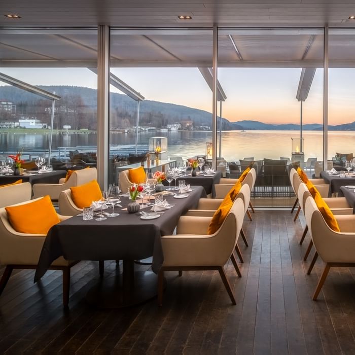 Elegant lakeside restaurant with beige chairs, orange cushions, and floor-to-ceiling windows at sunset at Falkensteiner Schlosshotel Velden