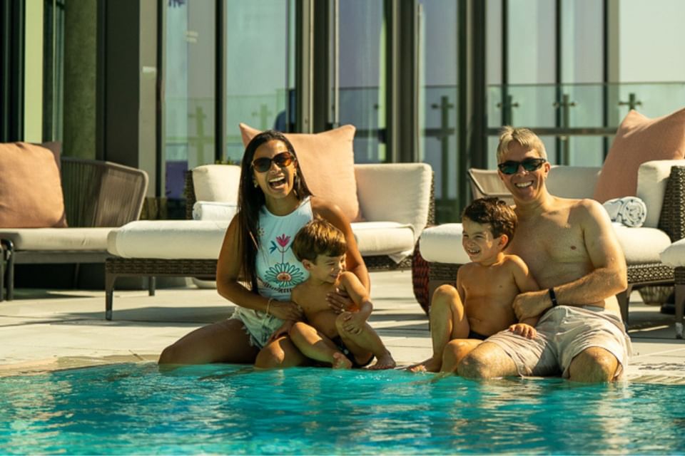Family sits at the pool edge beside cushioned loungers with pink pillows under heat lamps at Hotel X Toronto
