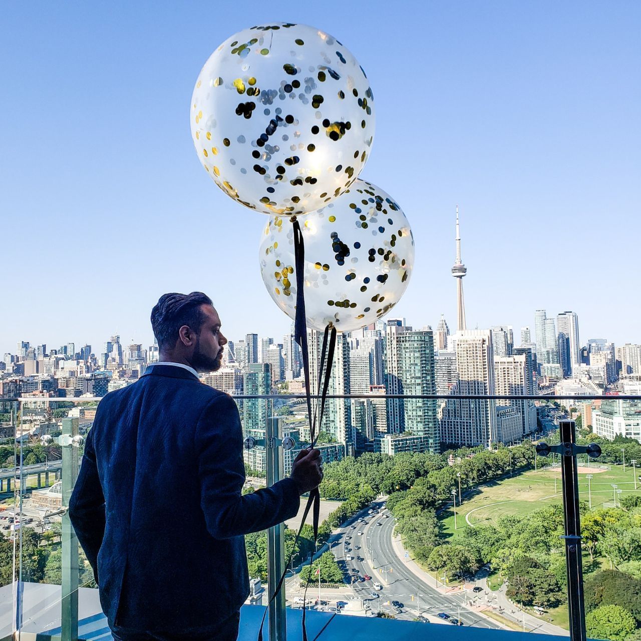 Man holding two large gold and black confetti balloons on a balcony overlooking a city.