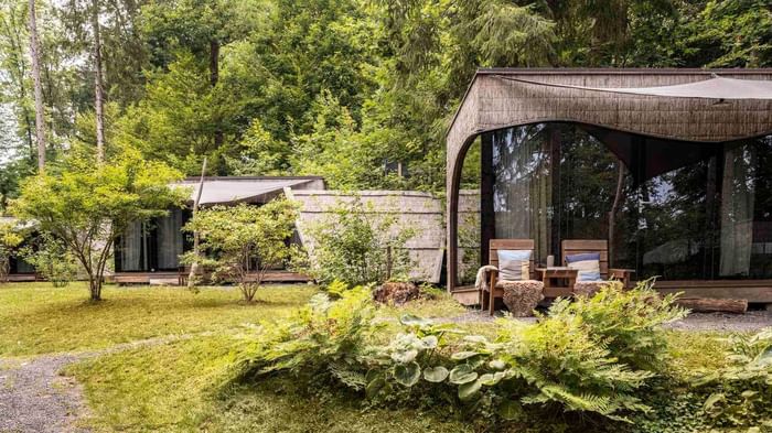 Two wooden chairs in front of glass-fronted cabins amidst lush greenery at Falkensteiner Premium Camping Lake Blaguš