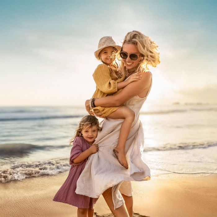 Eine Mutter trägt ihr Kind und umarmt ein weiteres Kind am Strand.