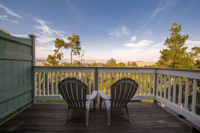 Two chairs on a wooden deck with railings, overlooking trees and mountains under a partly cloudy sky.