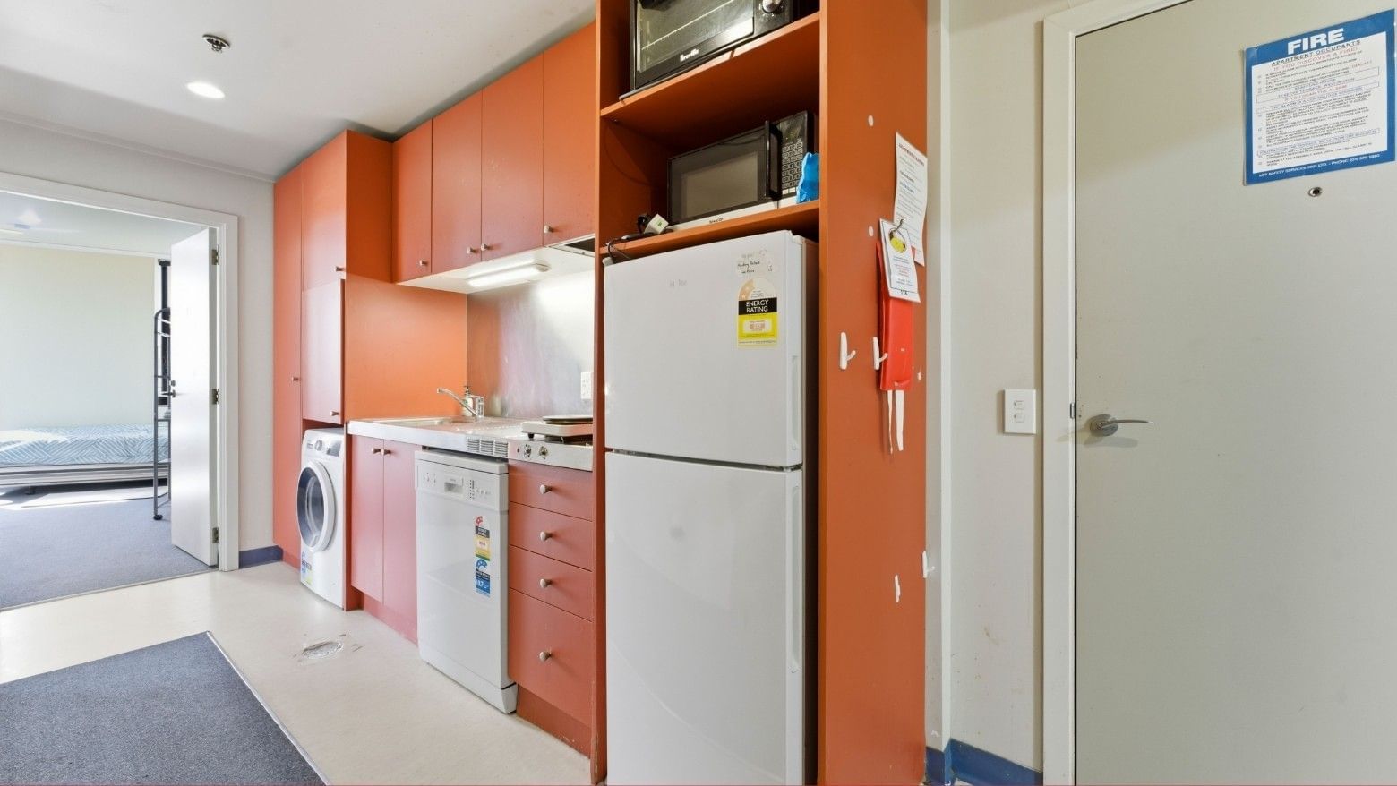 Kitchen area with orange cabinets, white appliances, and a bedroom in the background.