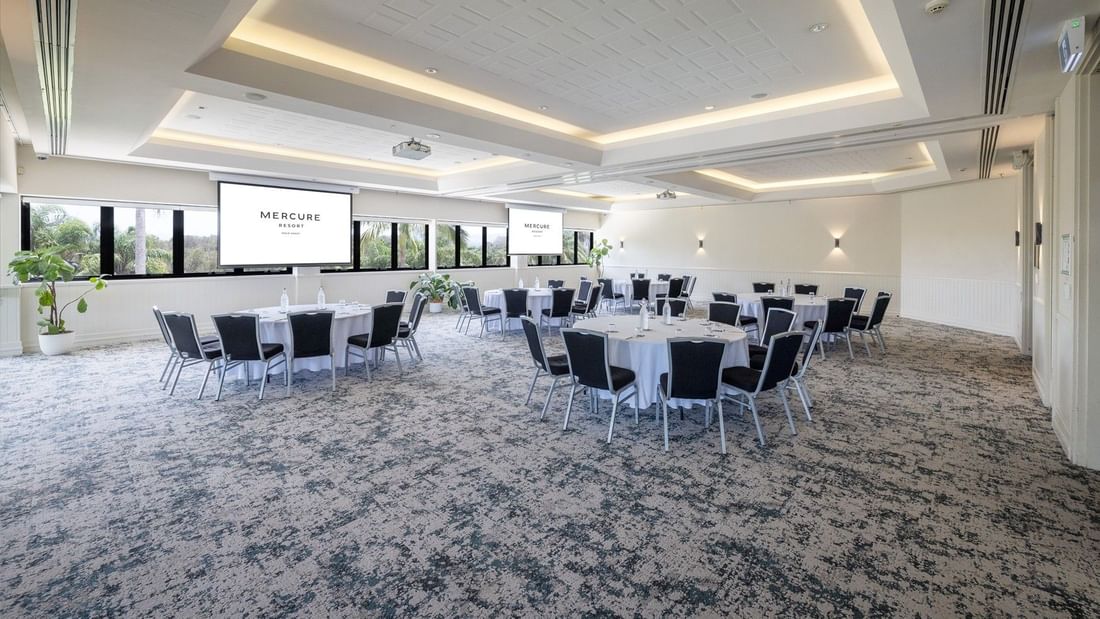 Large conference room with round tables, black chairs, two large screens, and potted plants by windows.