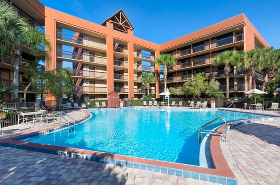 Orange multi-story hotel buildings with balconies around a pool with chairs and palm trees at Rosen Inn Lake Buena Vista