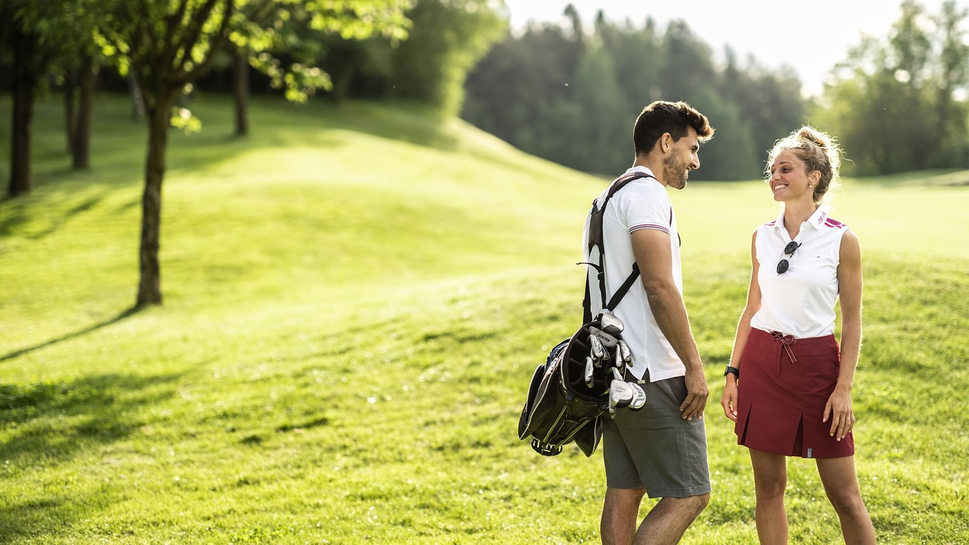 Man and woman in golf attire standing on a grassy field, smiling and carrying golf equipment.