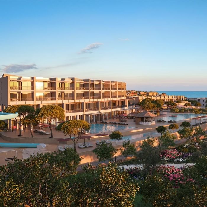Blick auf ein Resort mit Pool, Gebäuden, Bäumen und Meer im Hintergrund bei Sonnenuntergang.