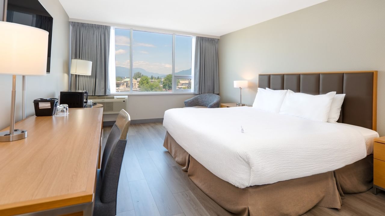 Hotel room with large king bed, desk, chairs, TV and large window with city view.