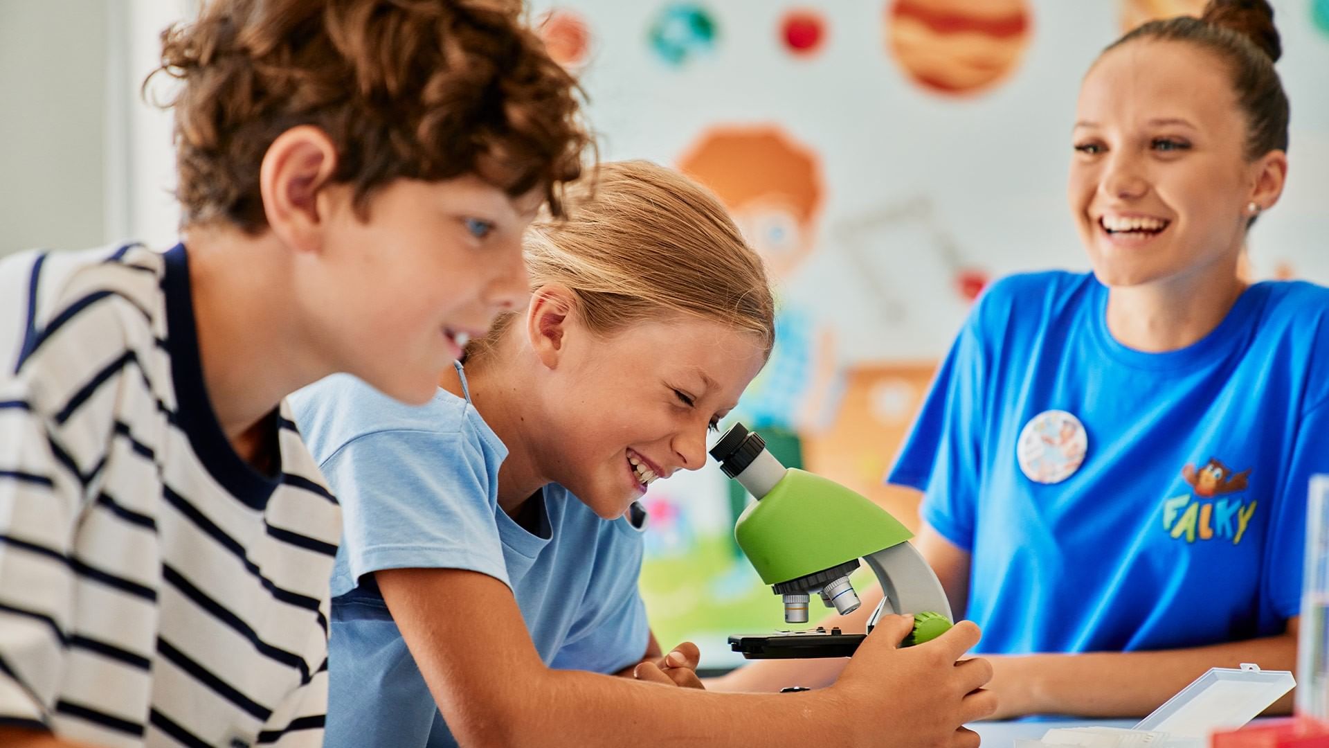Zwei Kinder und ein Betreuer in einem Klassenzimmer, die durch ein Mikroskop schauen.