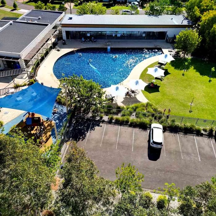 Aerial view of a pool with people swimming, surrounded by loungers, grass, and a parking lot at Mercure Kooindah Waters
