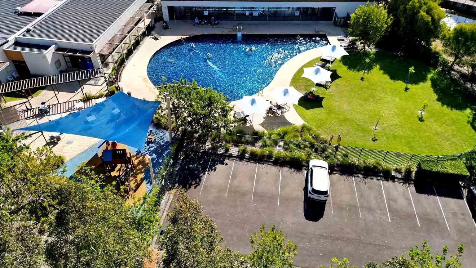 Aerial view of a pool with people swimming, surrounded by loungers, grass, and a parking lot at Mercure Kooindah Waters