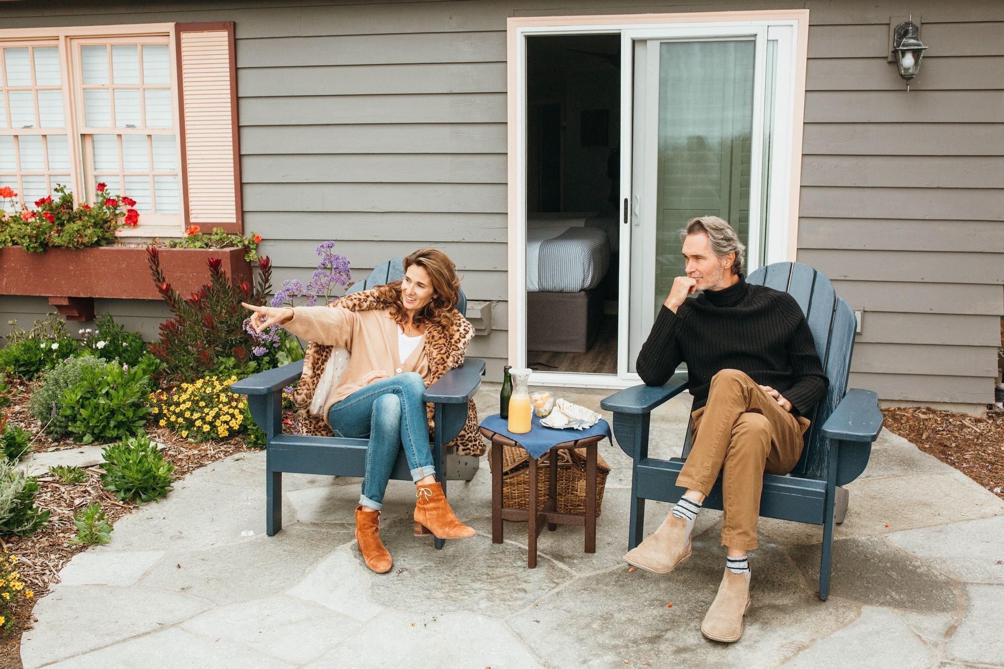 A couple sits on blue Adirondack chairs on a stone patio in front of a gray house with a garden.
