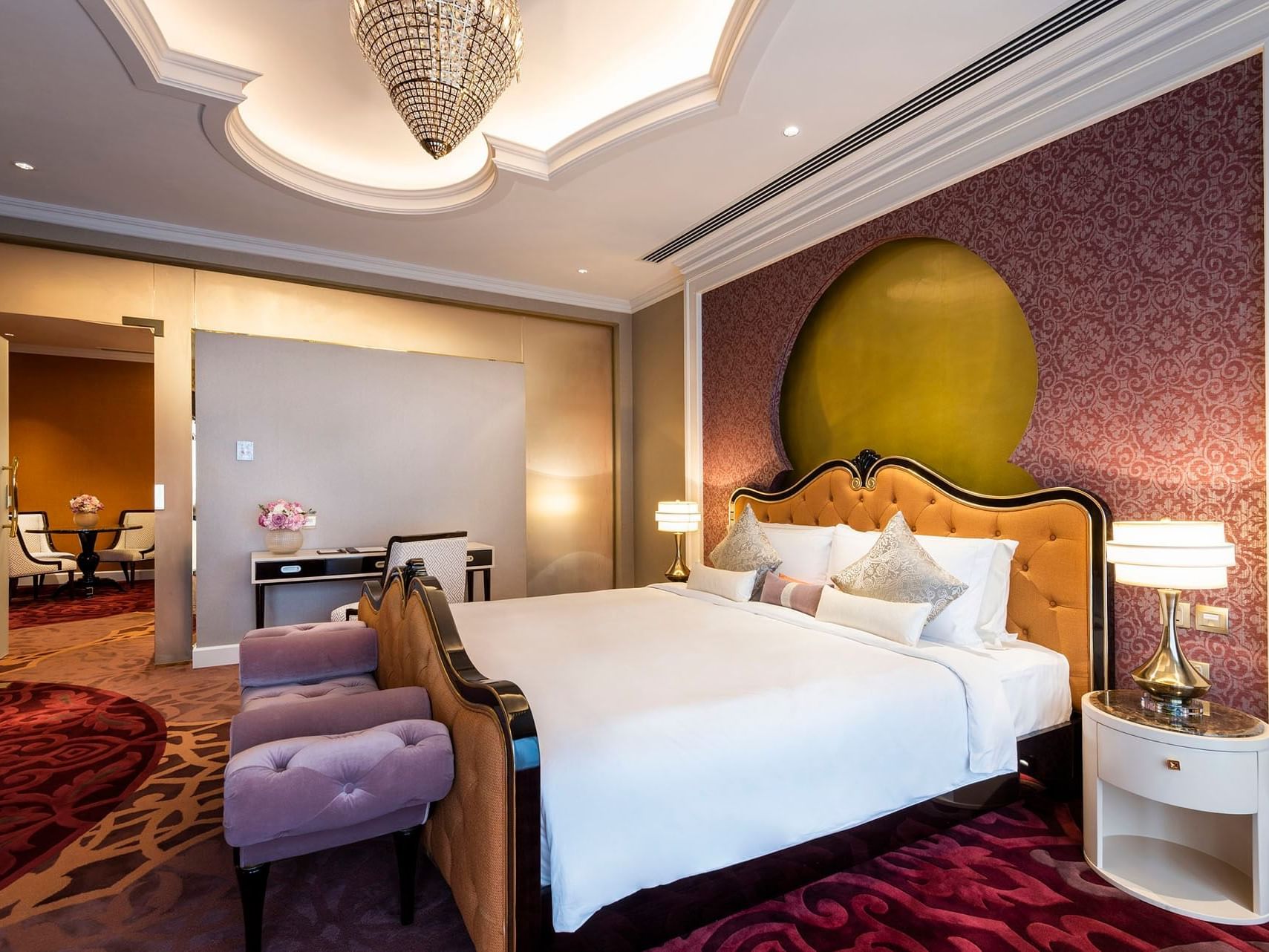 Elegant bedroom with large bed, purple chairs, patterned rug, and ornate headboard with chandelier and lamps.