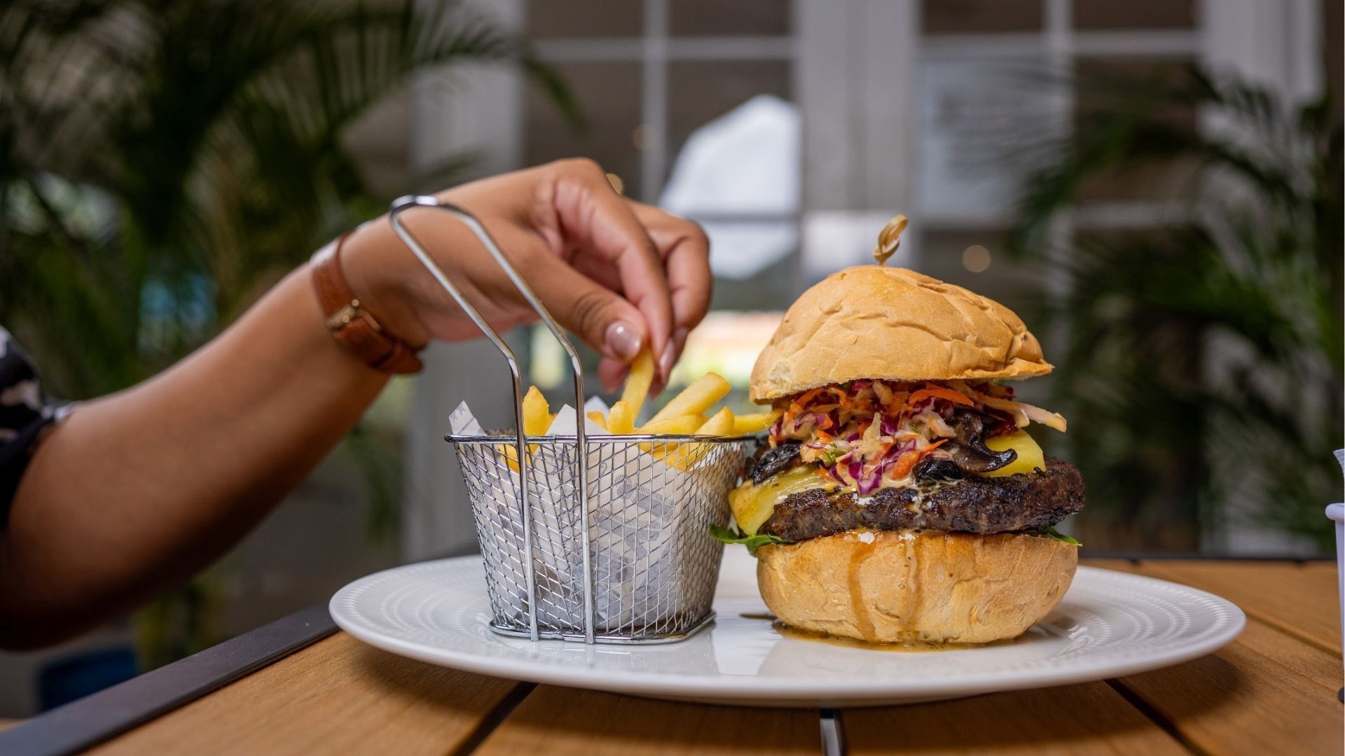 Gourmet burger on a plate by a basket of fries under a canopy of green plants in The Rhum Stop Restaurant at Time Out Hotel