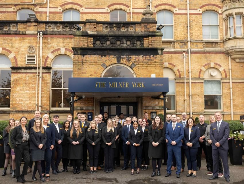 A large group of smiling staff in black and gold uniforms stands outside the entrance of The Milner York hotel in England