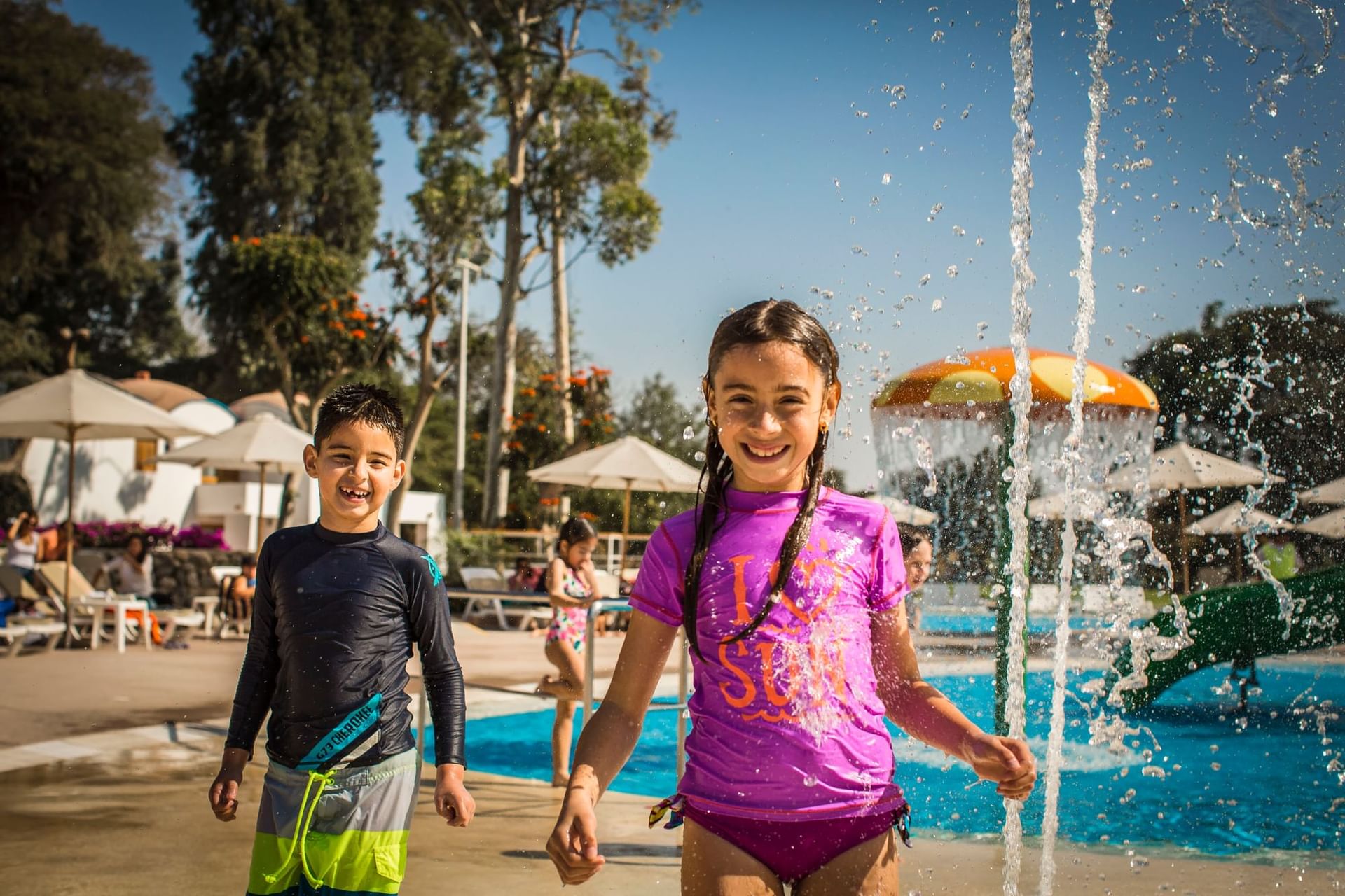 Two children play in a sunny pool area with umbrellas and greenery at Las Dunas Resort Ica Peru Hotels