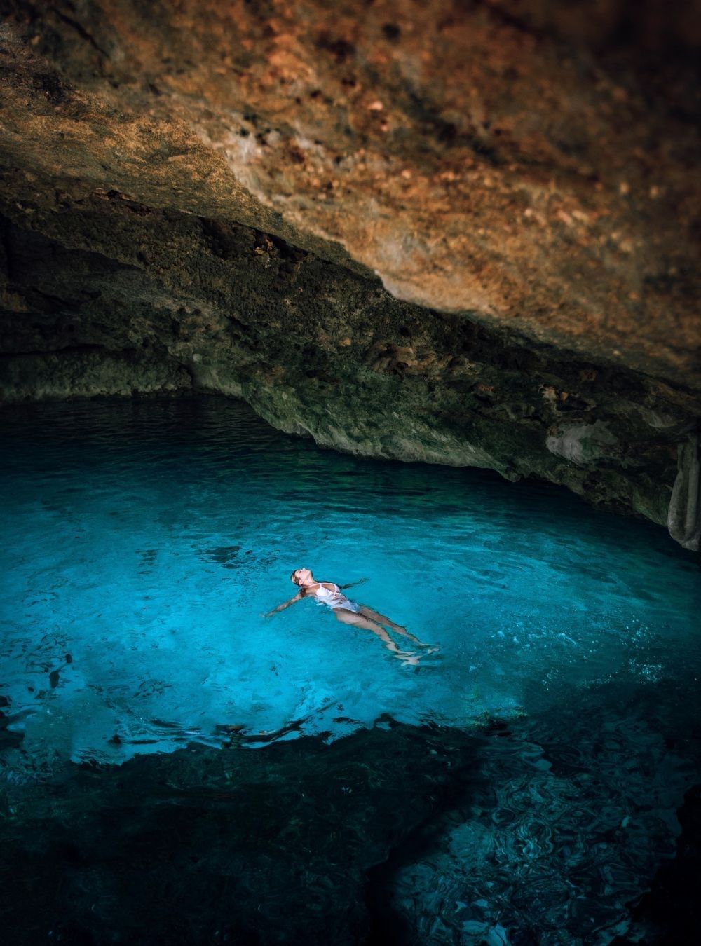 Woman floating in a turquoise underground cenote near Camino Real Pedregal Mexico, under a rocky cave ceiling