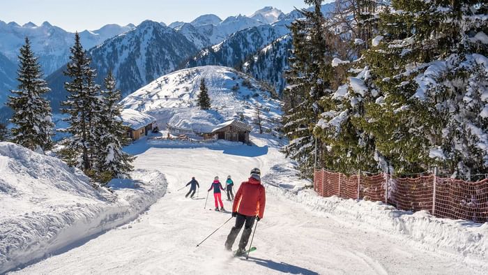 Vier Skifahrer fahren eine schneebedeckte Piste hinunter, umgeben von schneebedeckten Bäumen und Bergen.