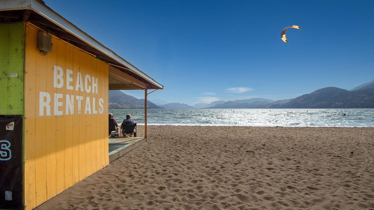 Two people sit outside a yellow hut on a beach, with kite surfing in the background.
