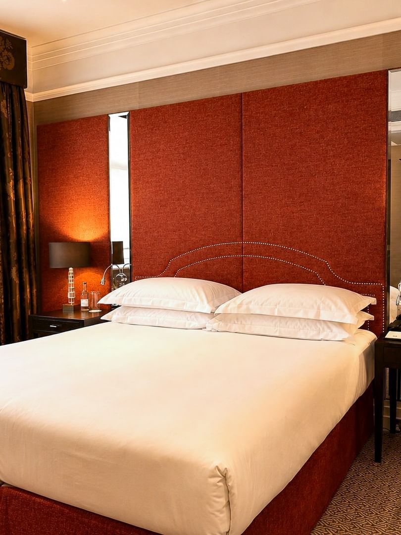 White bed by a dark nightstand under a red headboard near brown floral curtains at Warwick Hotels & Resorts