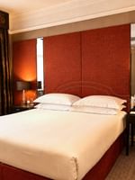 White bed by a dark nightstand under a red headboard near brown floral curtains at Warwick Hotels & Resorts