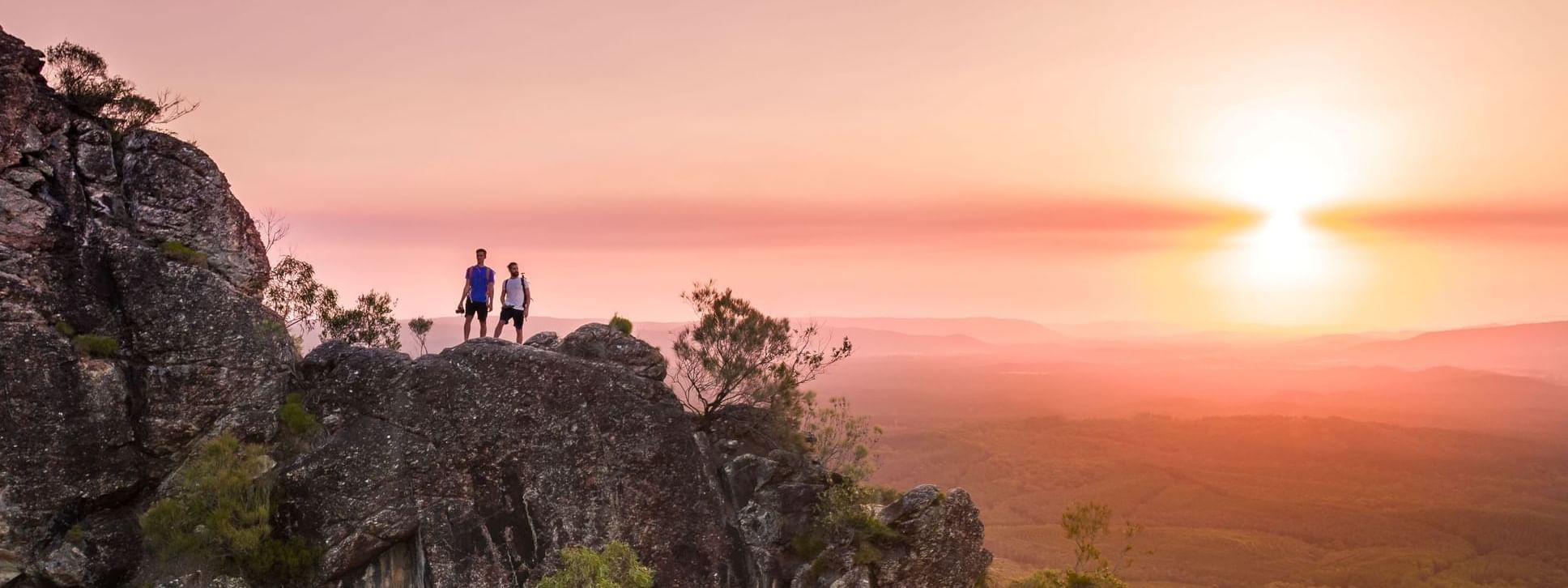 Two hikers on a rocky outcrop silhouetted by a vibrant sunset over a valley and mountains near Novotel Sunshine Coast Resort