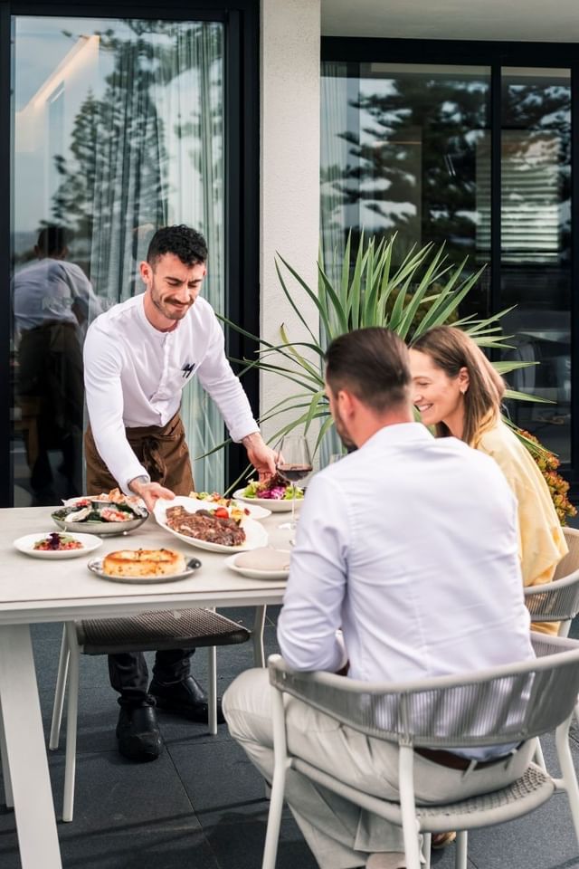 Waiter serving food to a couple at an outdoor dining table surrounding the Elysium Manly, a fine 5-star hotel in Manly