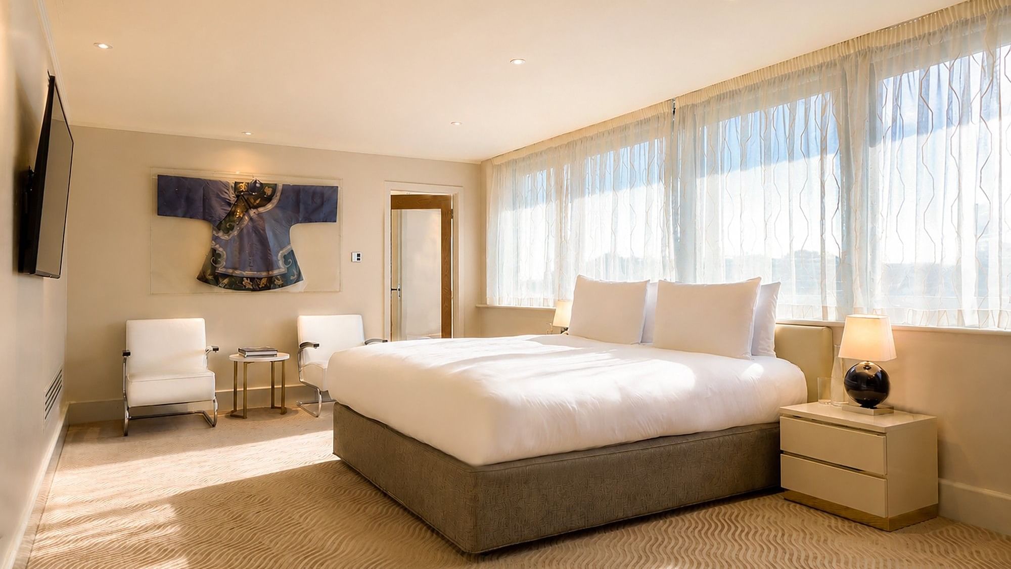 Bright Penthouse Suite bedroom with king bed, white armchairs, decorative wall art, windows at The May Fair Hotel, London.