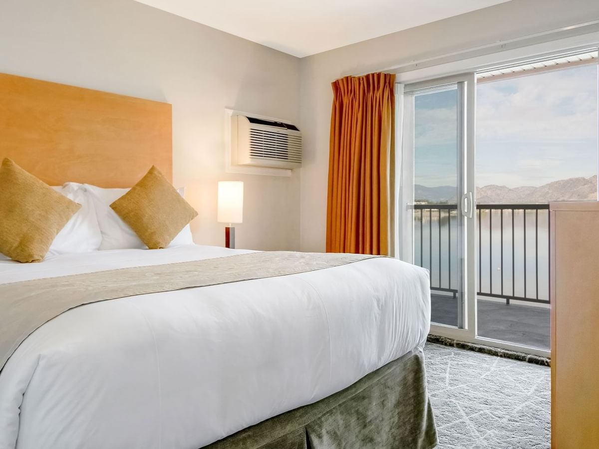 Queen bed in a bright room with a balcony overlooking serene lake scenery.