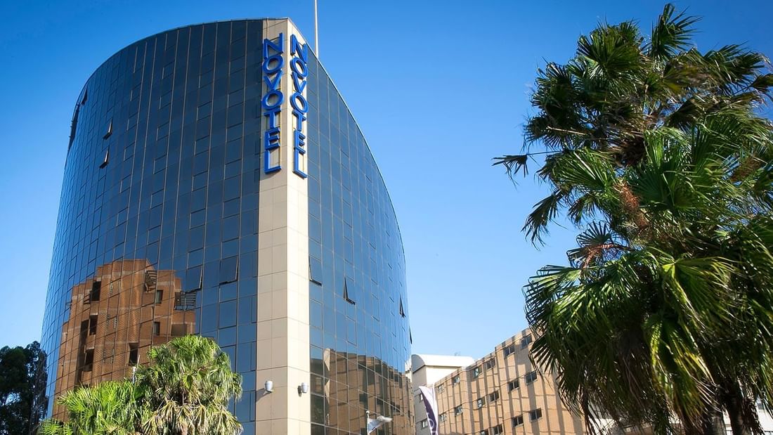 Modern Novotel Parramatta hotel building with palm trees under a clear blue sky.