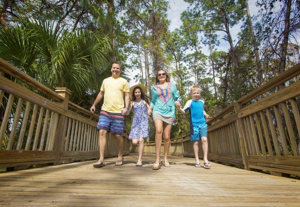 A family of four in summery attire walk along a wooden boardwalk toward the camera. 